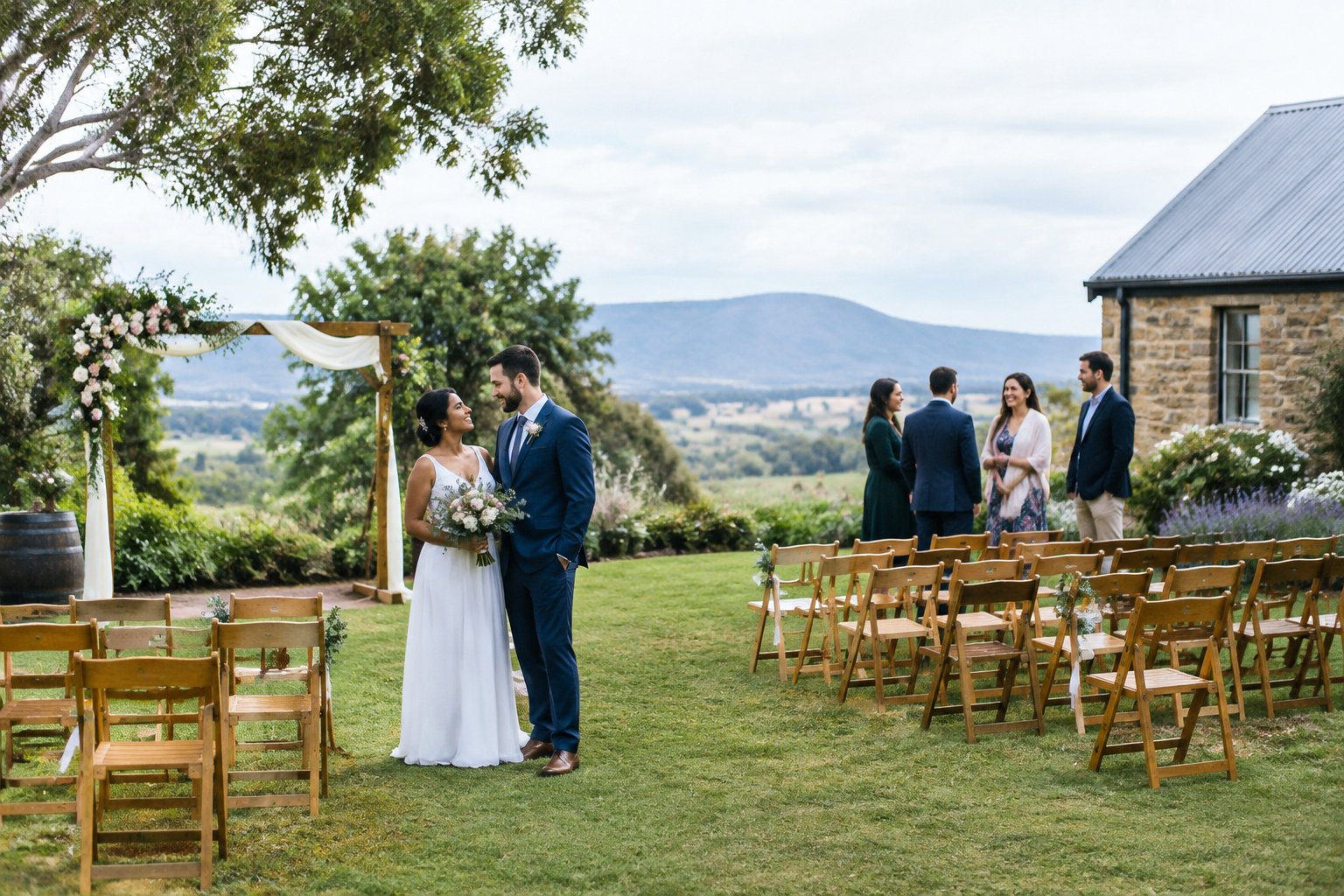 A couple stands beside a simple wedding ceremony setup in a welcoming outdoor venue with guests nearby.