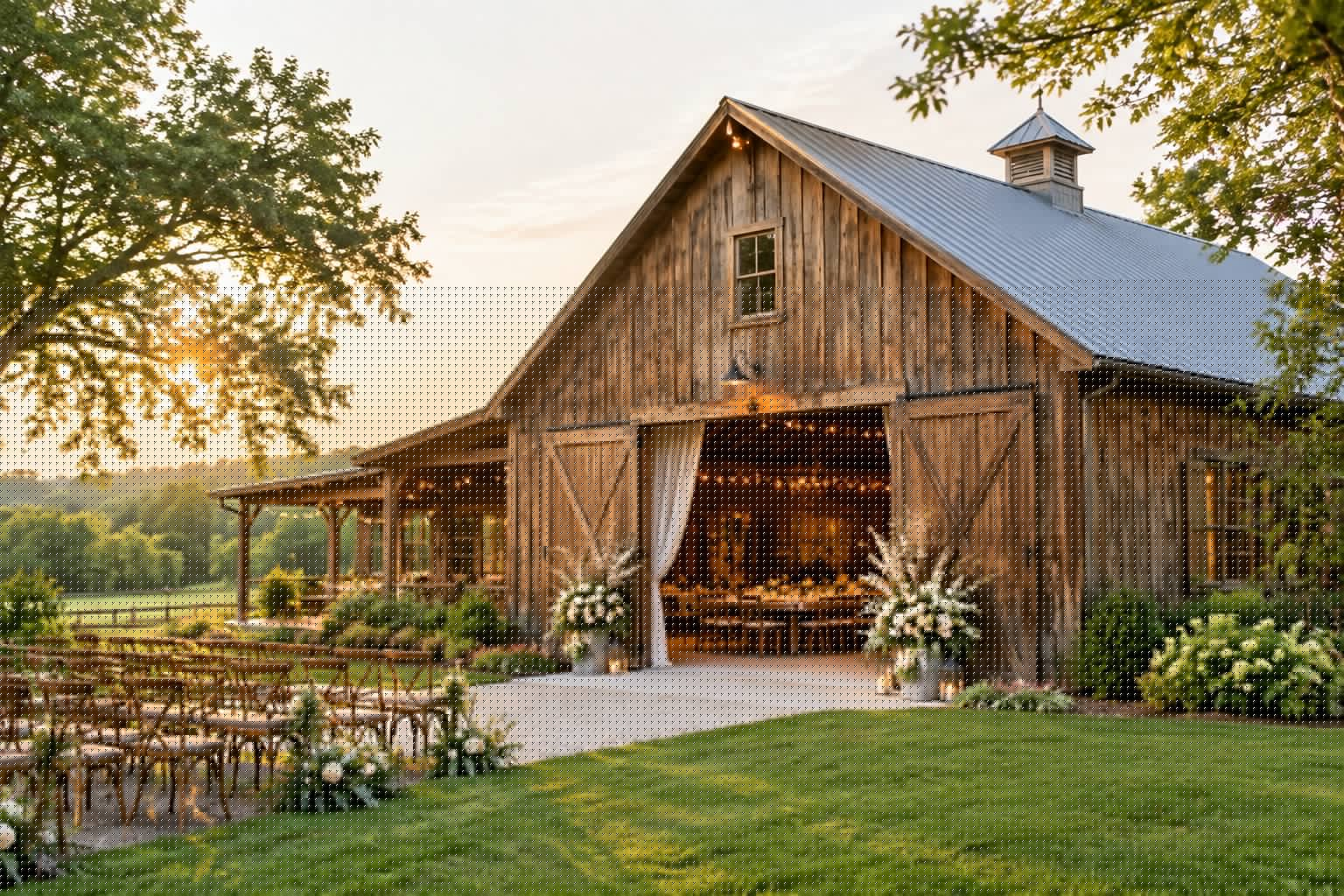 A rustic barn wedding venue surrounded by green countryside, with flowers, outdoor seating, and warm string lights.