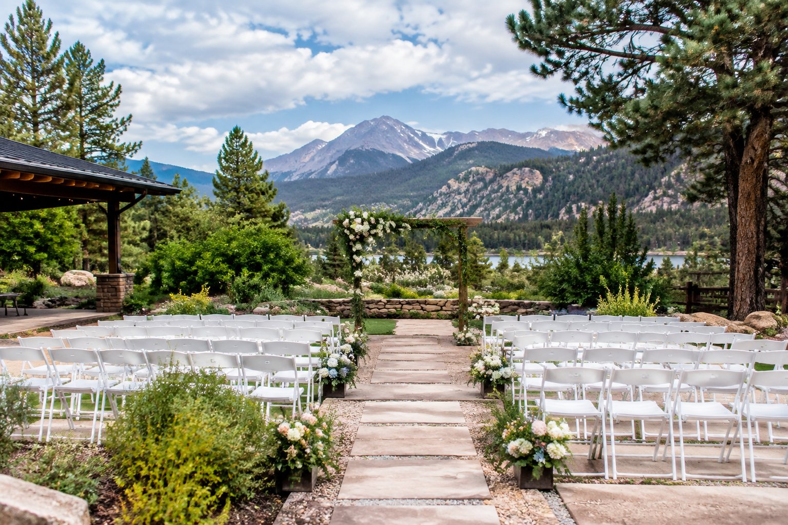 An outdoor garden wedding ceremony setup in Estes Park with white chairs, flowers, mountain scenery, and a covered area nearby.