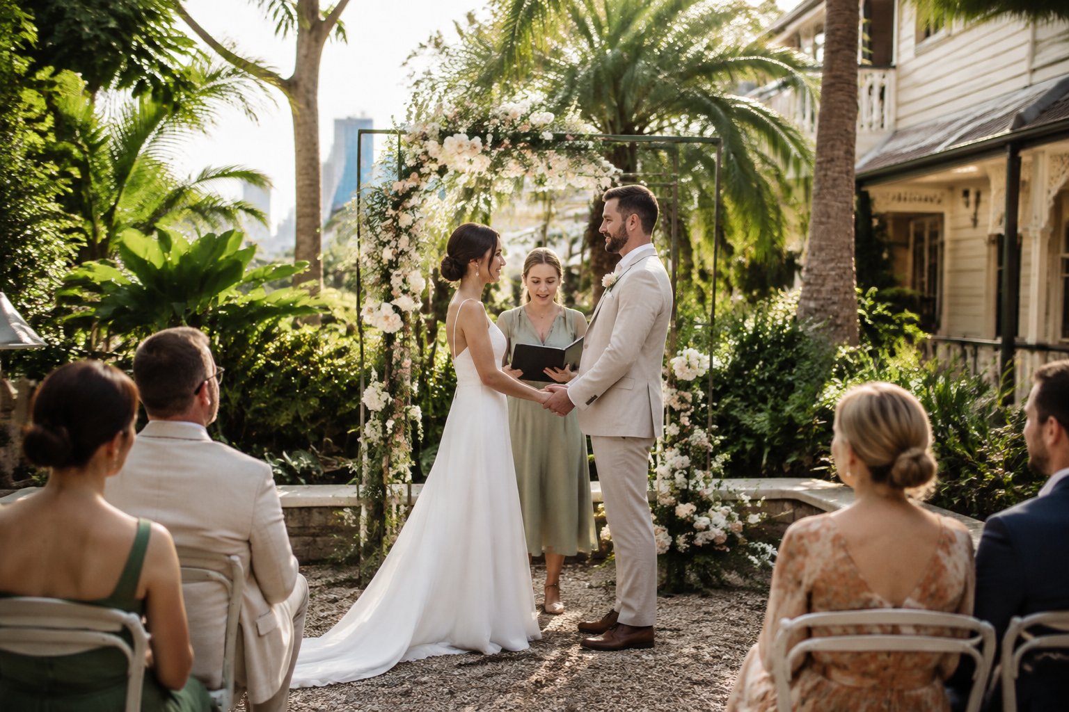 A couple exchanges vows beneath a floral arch in a garden courtyard as a small group of guests watches.