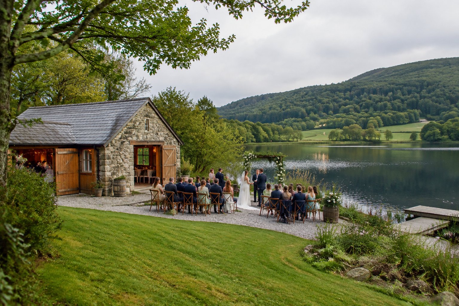 A small wedding gathering beside a lakeside barn surrounded by green woodland.
