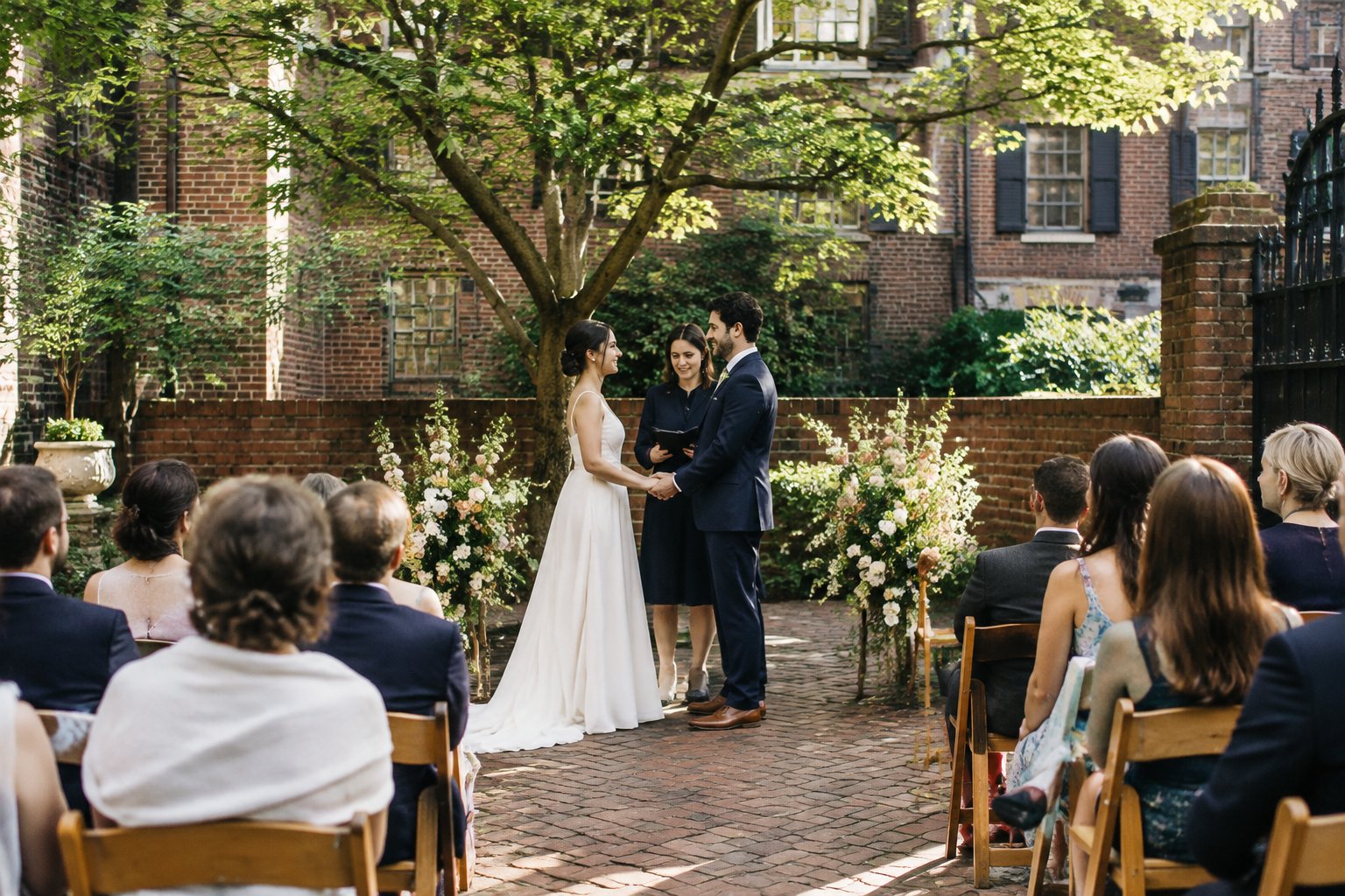 A couple stands together during a small wedding ceremony in a brick courtyard as guests watch.