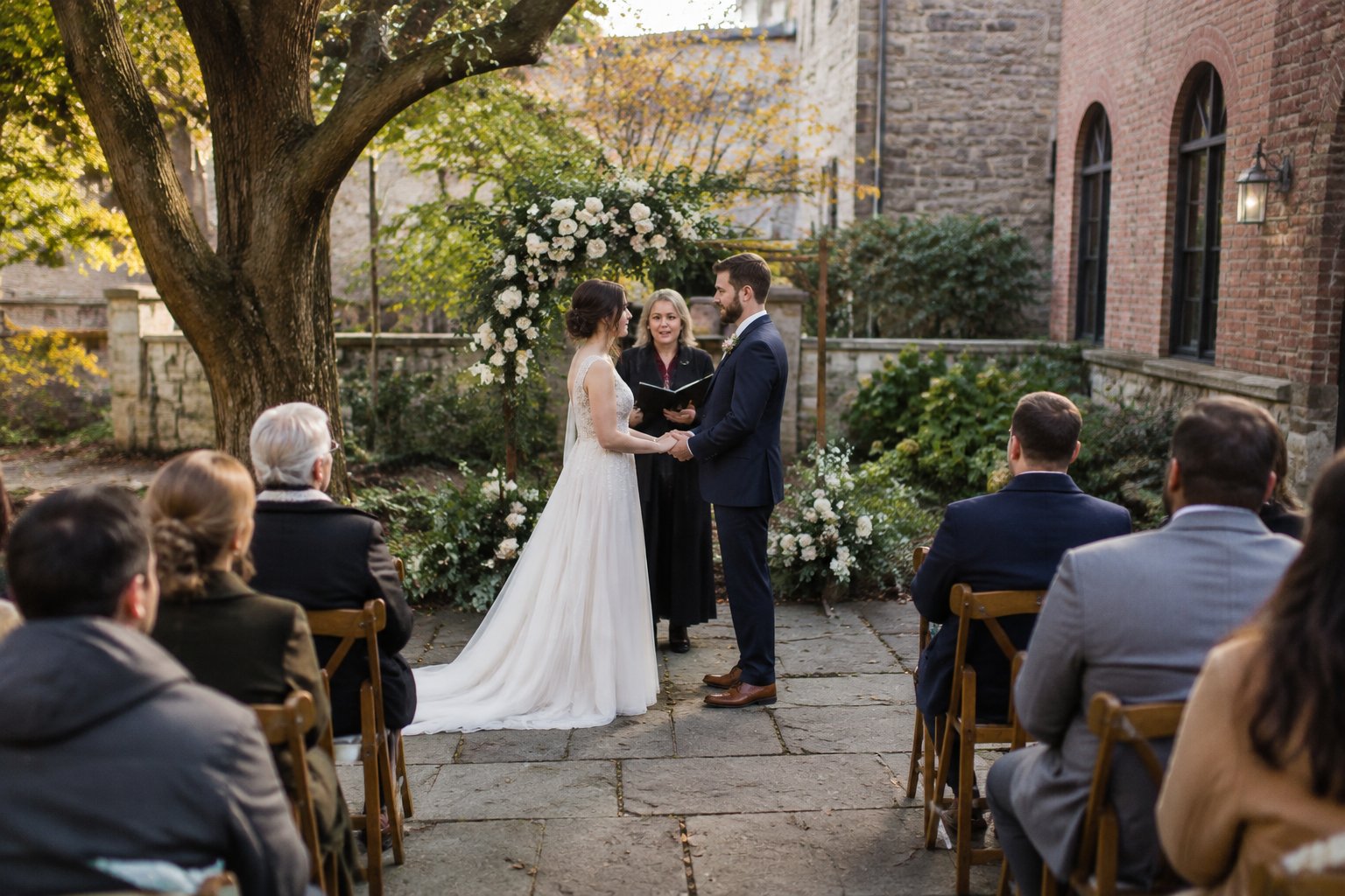 A couple exchanges vows beneath a floral arch as guests watch in a garden courtyard.