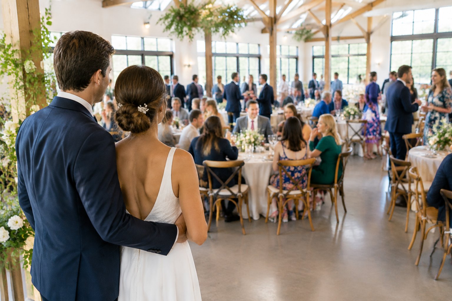 A couple looks across a wedding reception as guests mingle around dining tables.