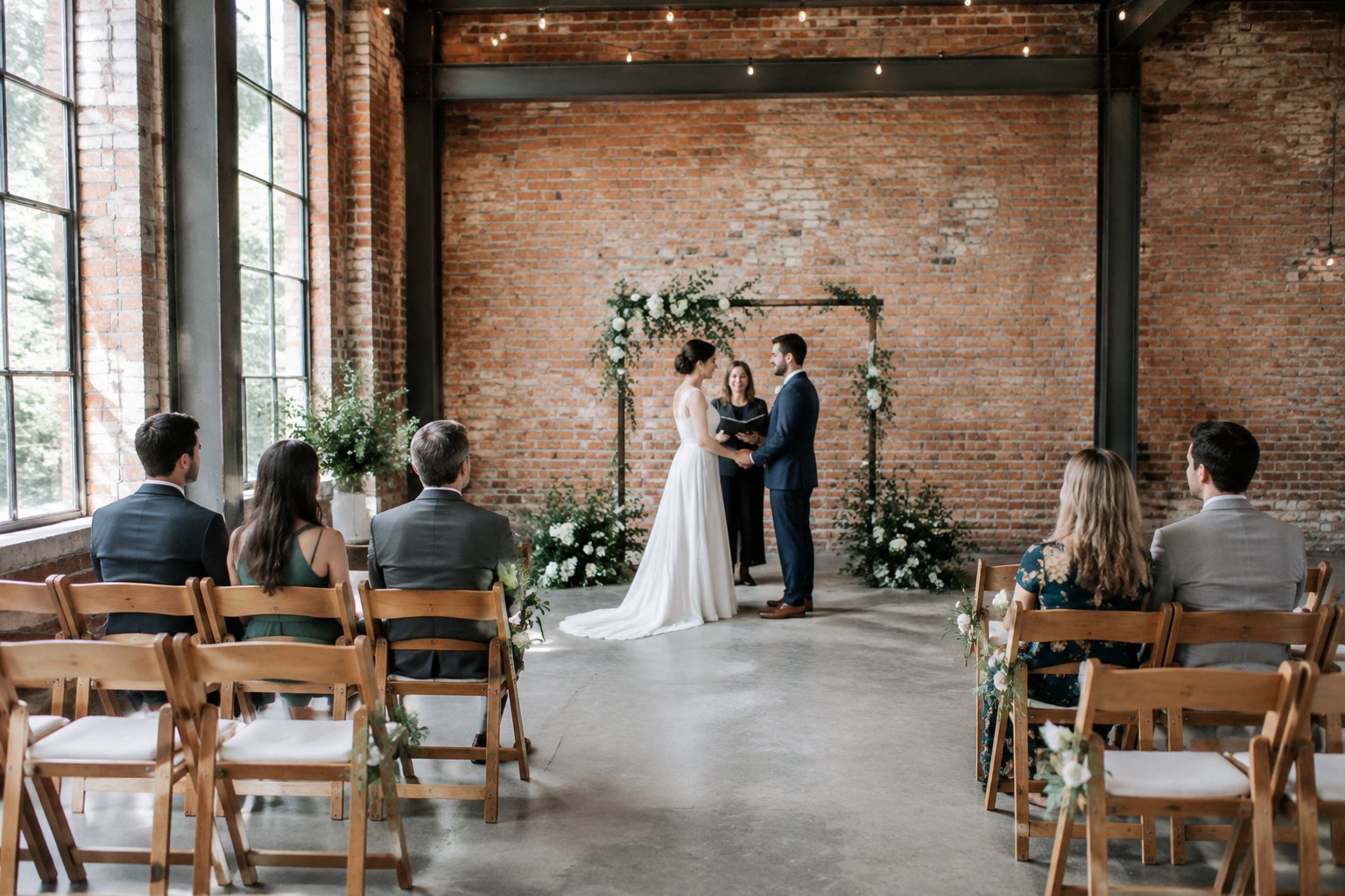 A couple stands at the front of a small wedding ceremony in a warehouse.