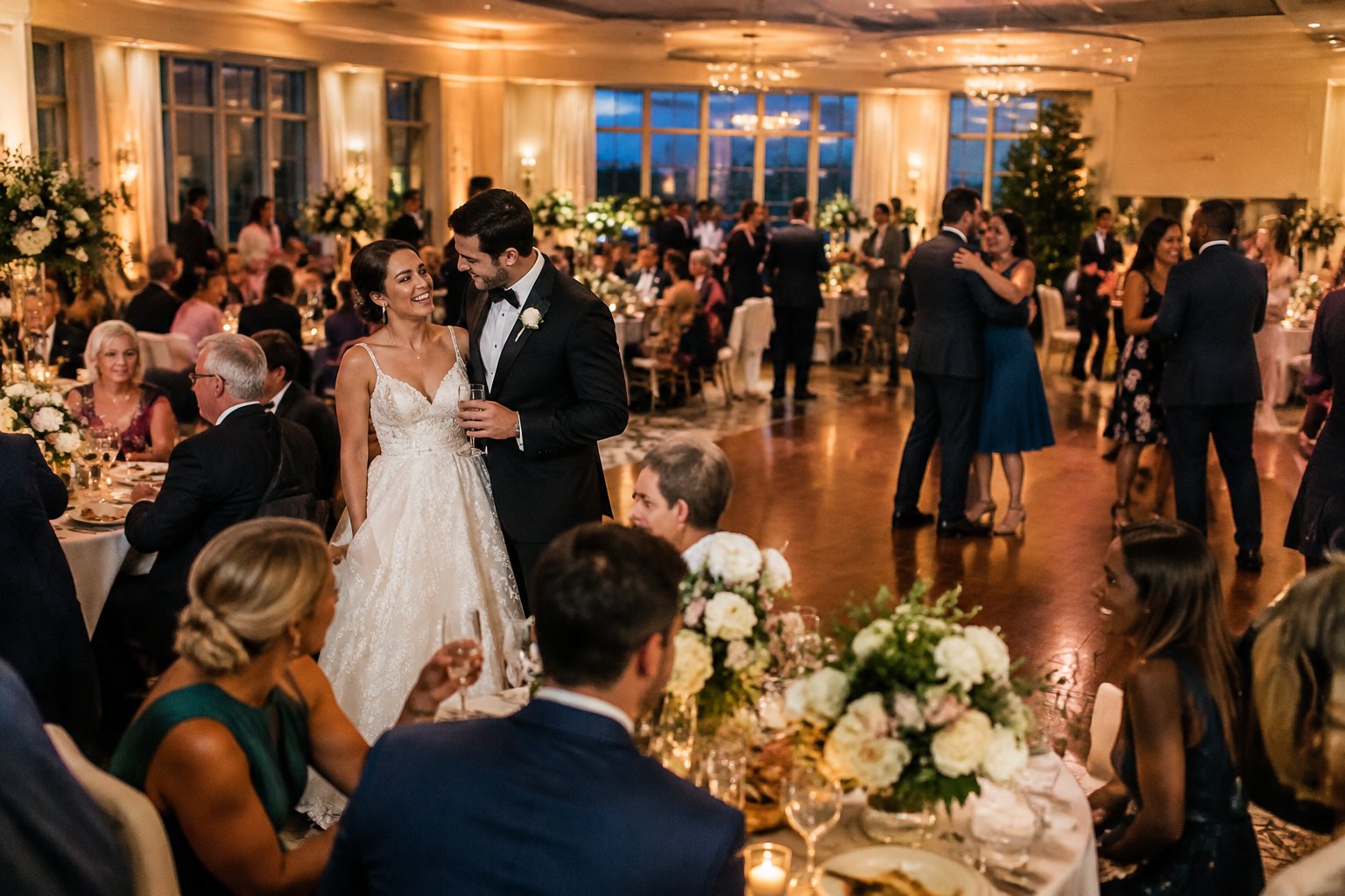 A newlywed couple celebrates with guests at a warmly lit wedding reception.