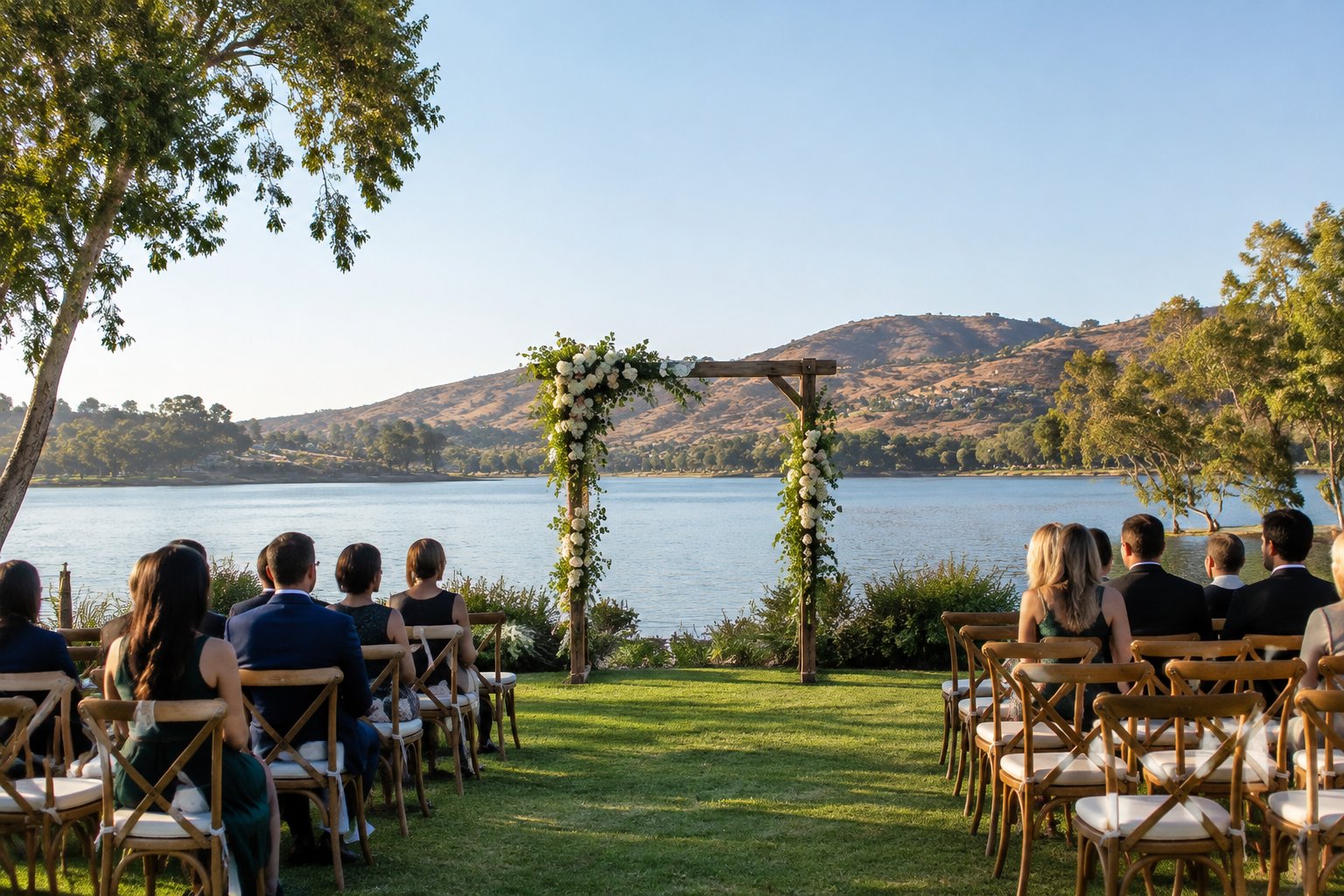 An outdoor wedding ceremony beside a lake, with chairs on a lawn and hills in the distance.